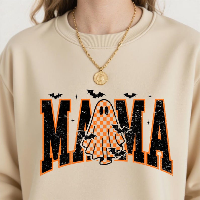 K&ouml;nnte beinhalten: Beiges Sweatshirt mit dem Wort "MAMA" in verwitterten schwarzen und orangefarbenen Buchstaben. Eine karierte Geistergrafik befindet sich in der Mitte, umgeben von Flederm&auml;usen. Das Sweatshirt ist ein Halloween-Design.