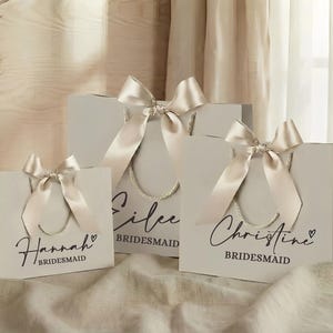 Puede incluir: Tres bolsas de regalo de color gris claro con lazos de cinta de sat&eacute;n marfil. Cada bolsa tiene un nombre en escritura negra: Hannah, Eilee y Christine. La palabra "Bridesmaid" est&aacute; debajo de cada nombre. Las bolsas tienen asas de cuerda.