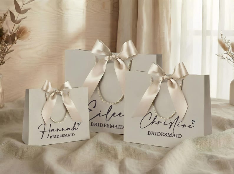 Puede incluir: Tres bolsas de regalo de color gris claro con lazos de cinta de sat&eacute;n marfil. Cada bolsa tiene un nombre en escritura negra: Hannah, Eilee y Christine. La palabra "Bridesmaid" est&aacute; debajo de cada nombre. Las bolsas tienen asas de cuerda.