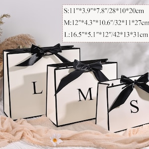 Puede incluir: Tres bolsas de regalo color crema con ribetes y lazos negros, etiquetadas con S, M y L. La bolsa m&aacute;s peque&ntilde;a mide 28 cm de ancho, 10 cm de profundidad y 20 cm de alto. La bolsa mediana mide 32 cm de ancho, 11 cm de profundidad y 27 cm de alto. La bolsa m&aacute;s grande mide 42 cm de ancho, 13 cm de profundidad y 31 cm de alto.