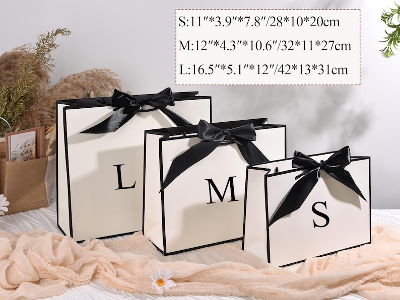 Puede incluir: Tres bolsas de regalo color crema con ribetes y lazos negros, etiquetadas con S, M y L. La bolsa m&aacute;s peque&ntilde;a mide 28 cm de ancho, 10 cm de profundidad y 20 cm de alto. La bolsa mediana mide 32 cm de ancho, 11 cm de profundidad y 27 cm de alto. La bolsa m&aacute;s grande mide 42 cm de ancho, 13 cm de profundidad y 31 cm de alto.