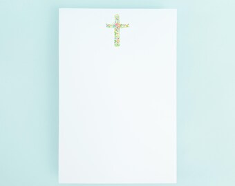May include: A white notepad with a floral cross design at the top. The cross is composed of small, colorful flowers and leaves. The background is a light blue.