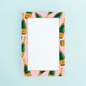 May include: A pink notepad with a gold border and a pattern of green champagne bottles with gold foil labels. The notepad has a large white space for writing. The background is a light blue.