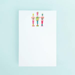 May include: Three watercolor nutcracker illustrations in pink, green, and blue adorn a white notepad. Each nutcracker has a different hat and uniform, with candy cane and bow details. The notepad is set against a light blue background.
