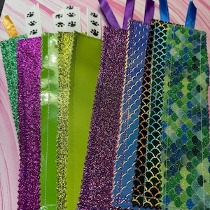 May include: A collection of rectangular bookmarks featuring various textures and patterns, including purple glitter, lime green vinyl, and iridescent blue and green scale designs. Each bookmark is topped with a small ribbon in matching colors.