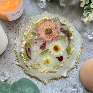 Puede incluir: Un posavasos redondo de resina blanquecina con borde dorado. El posavasos contiene flores secas, incluyendo margaritas y una gran rosa rosa. Un artículo decorativo.