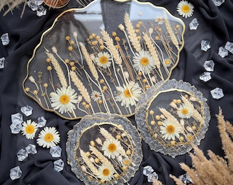 May include: Three clear resin trays with gold leaf accents and pressed dried flowers, including daisies and wheat, on a black fabric background.