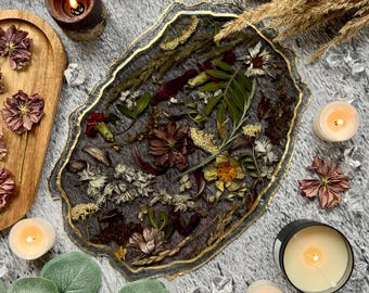 May include: A decorative resin tray with dried flowers and gold trim. The tray is surrounded by lit candles, faux greenery, and decorative crystals. A wooden tray with dried flowers is also present.