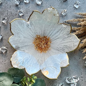Peut inclure: Plat en forme de fleur en résine blanche avec un bord doré et des paillettes dorées au centre.