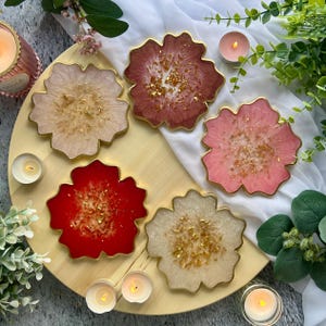 May include: A set of five flower-shaped resin coasters with gold leaf accents. The coasters feature a smooth, glossy finish with scalloped edges in shades of cream, deep red, dusty rose, light pink, and off-white, each with a gold-toned rim.