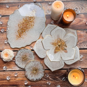 May include: Set of four white resin coasters with gold glitter accents. The coasters are shaped like a flower and a freeform shape. The coasters are displayed on a wooden surface with candles.