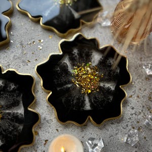 May include: Black flower-shaped coasters with gold edging and glitter centres. The coasters are resin and have a glossy finish. A lit candle and decorative crystals are also visible.