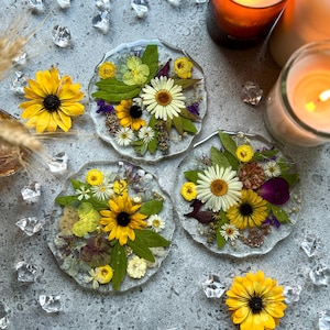 Puede incluir: Tres posavasos redondos de resina transparente con flores y hojas prensadas. Los posavasos presentan una variedad de flores amarillas, blancas y moradas, así como hojas verdes.