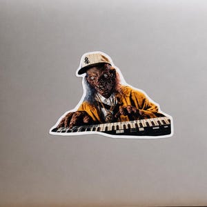 Peut inclure: Un autocollant représentant un zombie jouant du clavier. Le zombie porte une veste jaune, une casquette de baseball et une chaîne en or. Le clavier est noir et blanc. L'autocollant est placé sur un ordinateur portable.