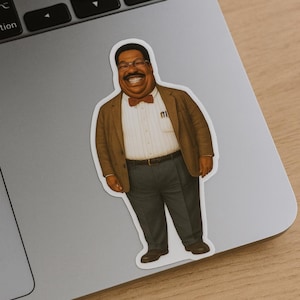 May include: A sticker featuring a smiling man in a brown blazer, bow tie, and glasses. He is wearing a white striped shirt and gray pants. The sticker is placed on a silver laptop.