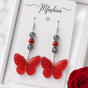 Peut inclure: Une paire de boucles d'oreilles pendantes avec des papillons rouges translucides suspendus à des crochets argentés. Chaque boucle comporte une perle rouge centrale entourée de deux perles grises craquelées sur une chaîne, sur un support Mfashion.