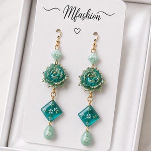 Peut inclure: Boucles d'oreilles pendantes avec crochet doré, perle losange vert clair à fleurs blanches, breloque fleur de lotus sarcelle, perle losange sarcelle à fleurs blanches et perle goutte vert clair. La carte présente l'inscription Mfashion.
