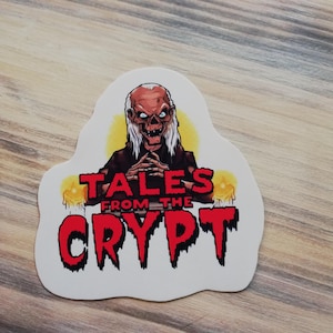 May include: A white sticker featuring a cartoon illustration of the Crypt Keeper from the TV show "Tales from the Crypt". The Crypt Keeper is depicted wearing a black robe and has a white beard. The text "Tales from the Crypt" is written in red lettering.