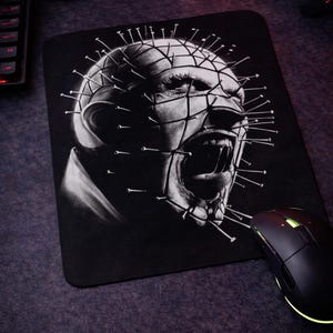 May include: Black mousepad featuring a grayscale illustration of a character with nails in their head and face. The character is screaming with their mouth open. A black computer mouse is on the right side of the mousepad. A skull is in the background.
