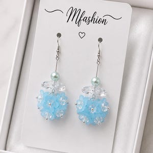 Peut inclure: Une paire de boucles d'oreilles pendantes sur un support blanc marqué Mfashion. Chaque boucle se compose d'un crochet argenté, d'une perle vert clair, d'un cristal facetté transparent et d'un groupe de perles en forme de fleurs bleu clair.