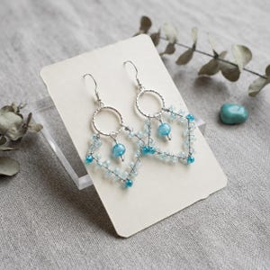 Peut inclure: Une paire de boucles d'oreilles pendantes argentées avec un anneau circulaire en haut, d'où pend une structure en fil métallique en forme de losange. Le cadre est orné de petites perles bleu clair et d'une perle bleue ronde et translucide.