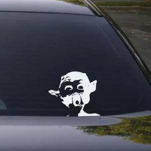 May include: A white vinyl sticker of a character from the movie 'Lord of the Rings' is on the back window of a black car. The character is a goblin with a large nose and big eyes.