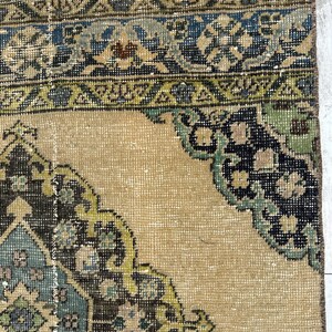 May include: A woven rug featuring a tan background with a central medallion and corner motifs in shades of dark brown, light blue, and sage green. The border displays geometric patterns in blue, green, and brown on a tan field.