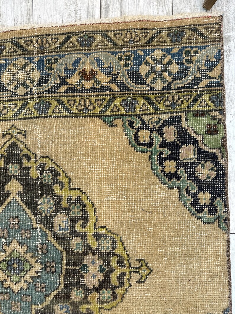 May include: A woven rug featuring a tan background with a central medallion and corner motifs in shades of dark brown, light blue, and sage green. The border displays geometric patterns in blue, green, and brown on a tan field.