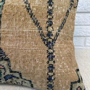May include: A rectangular beige cushion with a geometric pattern in dark blue, green, and pink. The cushion has a textured surface, and the design is reminiscent of a traditional rug. The cushion is likely for home decor.
