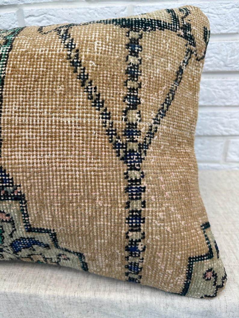 May include: A rectangular beige cushion with a geometric pattern in dark blue, green, and pink. The cushion has a textured surface, and the design is reminiscent of a traditional rug. The cushion is likely for home decor.