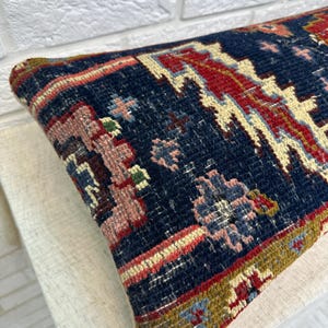 May include: A rectangular, patterned pillow with a navy blue background. The pillow features a detailed design with red, pink, and cream-colored geometric shapes and floral motifs. The edges are trimmed with a border of various colors.