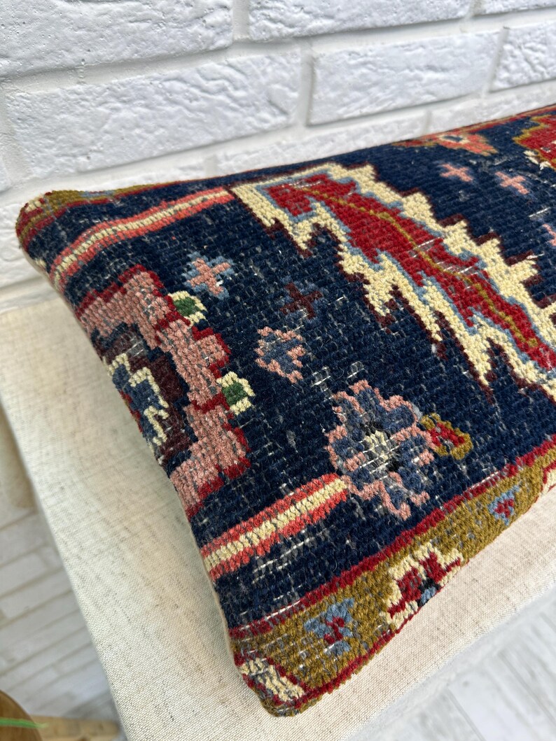 May include: A rectangular, patterned pillow with a navy blue background. The pillow features a detailed design with red, pink, and cream-colored geometric shapes and floral motifs. The edges are trimmed with a border of various colors.