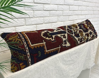 May include: A long, rectangular pillow with a patterned design. The pillow features a deep red background with navy blue, gold, and cream-colored geometric patterns. The pillow is placed on a white surface against a white brick wall.