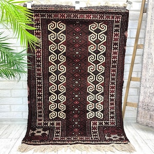 May include: A rectangular, dark brown rug with intricate geometric patterns in cream, black, and red. The rug features a central design of repeating scroll-like shapes and a fringed edge. The rug is displayed against a white brick wall.