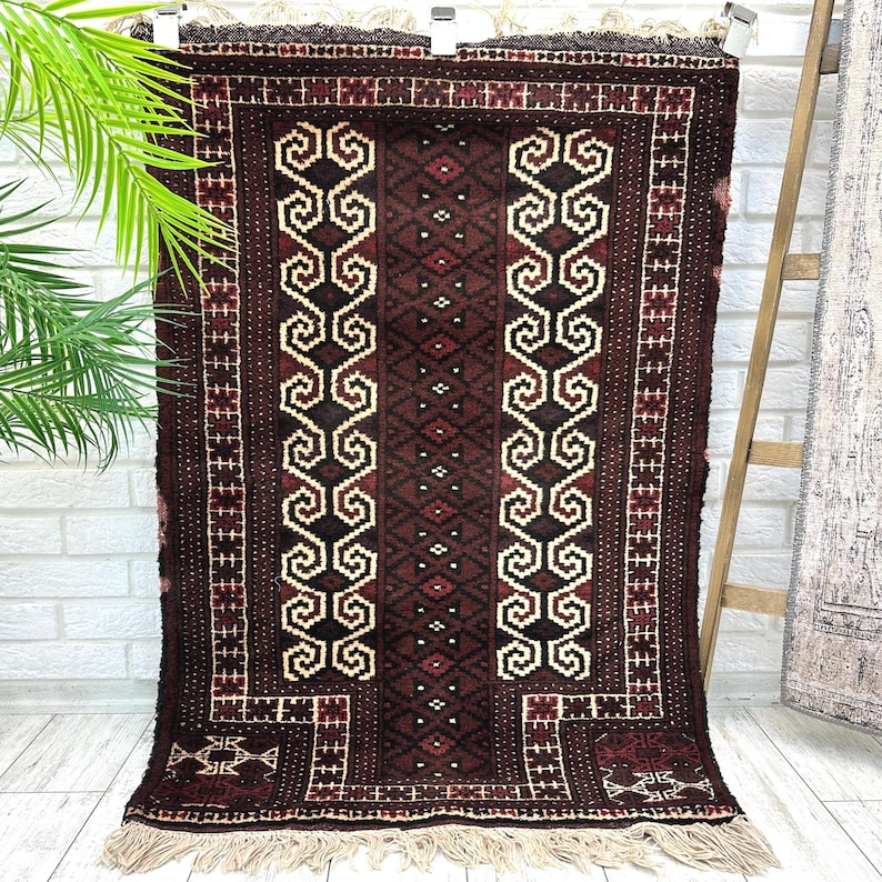 May include: A rectangular, dark brown rug with intricate geometric patterns in cream, black, and red. The rug features a central design of repeating scroll-like shapes and a fringed edge. The rug is displayed against a white brick wall.