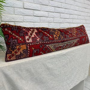 Puede incluir: Un cojín lumbar largo y rectangular con una base de color rojo intenso y patrones geométricos intrincados en tonos morado, naranja y crema. El tejido tiene una apariencia texturizada con detalles de flecos visibles a lo largo del borde superior.