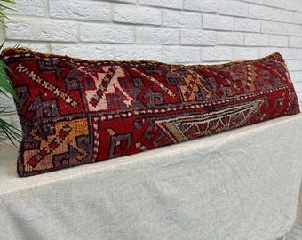 May include: A long, rectangular lumbar pillow featuring a deep red base color with intricate geometric patterns in shades of purple, orange, and cream. The textile has a textured, woven appearance with visible fringe detailing along the top edge.