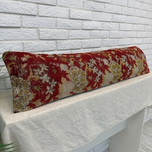 Könnte beinhalten: Ein langes, rechteckiges Lendenkissen mit einer tiefroten Grundfarbe und einem komplizierten Blumenmuster in Beige-, gedämpften Gelb- und Dunkelbrauntönen. Das Textil weist eine verwitterte, vintage-inspirierte Textur auf der gesamten Oberfläche auf.