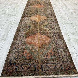 May include: A long, narrow runner rug featuring a repeating pattern of large, rounded geometric medallions in shades of orange, brown, green, and dark blue. The rug has a distressed, weathered texture with a central vertical line and fringe ends.