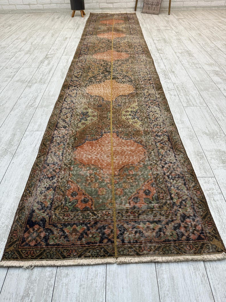 May include: A long, narrow runner rug featuring a repeating pattern of large, rounded geometric medallions in shades of orange, brown, green, and dark blue. The rug has a distressed, weathered texture with a central vertical line and fringe ends.