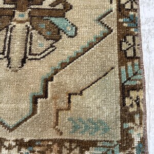 May include: Close-up of a beige and brown vintage rug with geometric patterns and fringe. The rug features a repeating design with brown and teal accents. The rug's texture is visible, showcasing its woven construction.