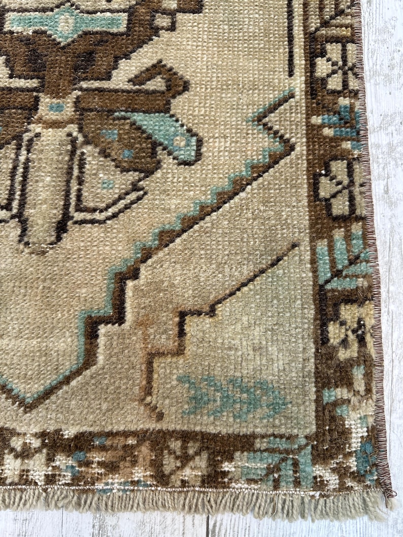 May include: Close-up of a beige and brown vintage rug with geometric patterns and fringe. The rug features a repeating design with brown and teal accents. The rug's texture is visible, showcasing its woven construction.