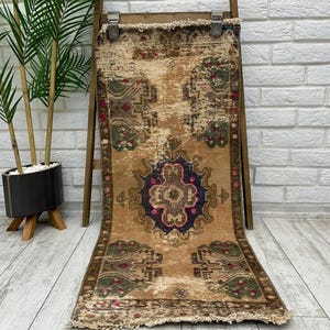May include: A vintage runner rug with a faded floral pattern in shades of brown, green, pink, and blue. The rug has a fringed edge and is displayed on a wooden stand.