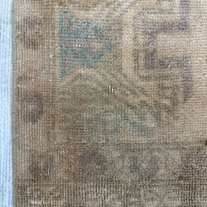 K&ouml;nnte beinhalten: Ein gewebter Teppich in einer Farbpalette aus Hellbraun und Beige mit einem verblassten geometrischen Muster. Das Design enth&auml;lt dezente Akzente in ged&auml;mpftem Gr&uuml;n und Braun und zeigt eine strukturierte Floroberfl&auml;che mit einer Kante an der Seite.