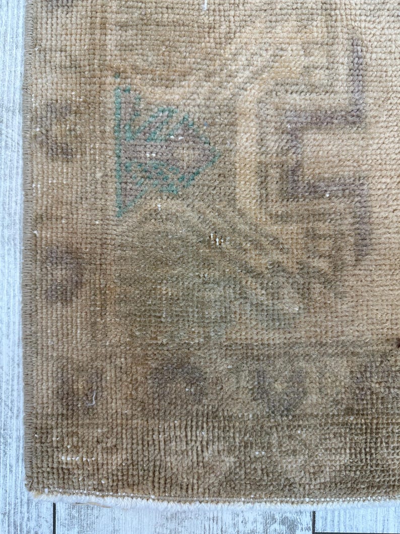 K&ouml;nnte beinhalten: Ein gewebter Teppich in einer Farbpalette aus Hellbraun und Beige mit einem verblassten geometrischen Muster. Das Design enth&auml;lt dezente Akzente in ged&auml;mpftem Gr&uuml;n und Braun und zeigt eine strukturierte Floroberfl&auml;che mit einer Kante an der Seite.