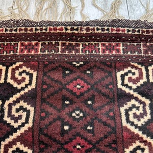 May include: Close-up of a section of a patterned rug. The rug features a deep burgundy and beige color palette with geometric and floral designs. The edges have a woven border and beige fringe.