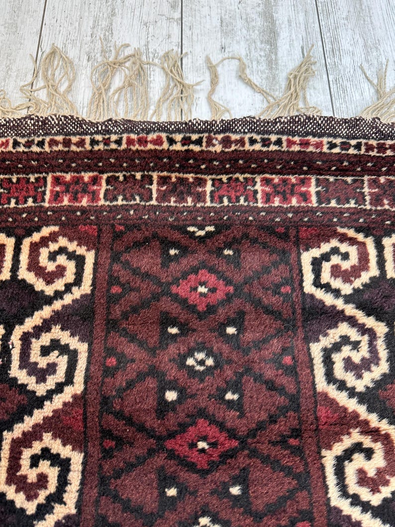 May include: Close-up of a section of a patterned rug. The rug features a deep burgundy and beige color palette with geometric and floral designs. The edges have a woven border and beige fringe.