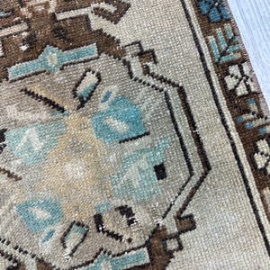 May include: Close-up of a beige and brown patterned rug with geometric shapes and floral accents. The rug features a border with a repeating design and is made of a woven material.