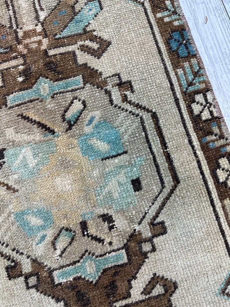 May include: Close-up of a beige and brown patterned rug with geometric shapes and floral accents. The rug features a border with a repeating design and is made of a woven material.