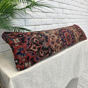 May include: A long, rectangular pillow with a detailed, vintage-style pattern. The pillow features a mix of red, blue, black, and beige hues, with floral and geometric designs. It rests on a white surface, with a white brick wall in the background.